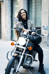 Glückliche Motorradfahrerin mit Helm auf einem Motorrad sitzend in der Stadt - JCMF01737