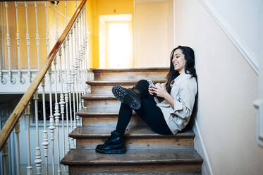 Happy young woman using smart phone while siting on staircase at home - JCMF01742