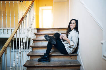 Smiling young woman with smart phone on steps at home - JCMF01743