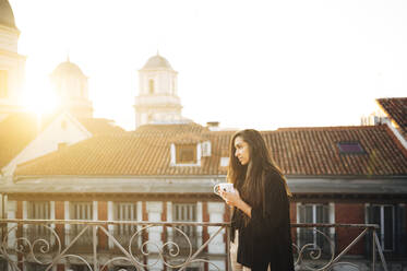 Young woman with coffee cup day dreaming while standing on rooftop in city - JCMF01745