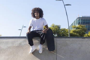 Lächelnder Mann mit Skateboard, der wegschaut, während er auf einer Rampe im Skateboardpark sitzt - PNAF00381