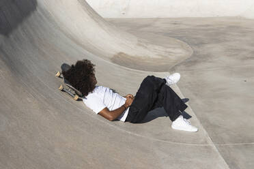 Sportsman with skateboard relaxing while lying on sports ramp at skateboard park - PNAF00394