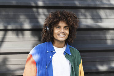 Smiling man wearing headphones listening music while standing against black wall - PNAF00396