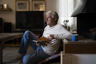 Nachdenklicher älterer Mann hält ein Buch, während er zu Hause auf dem Boden sitzt - AFVF07909