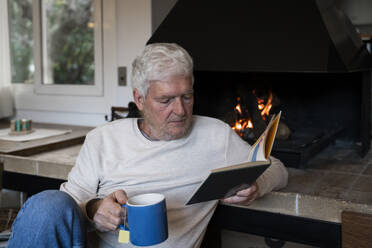 Älterer Mann mit Kaffeetasse, der ein Buch liest, während er zu Hause am Kamin sitzt - AFVF07910