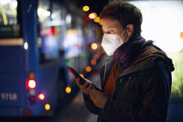 Frau mit Gesichtsmaske, die an einer Bushaltestelle steht und ein Mobiltelefon benutzt - MAMF01477
