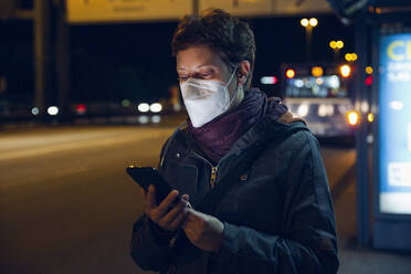 Woman wearing protective face mask using smart phone while standing at bus stop during COVID-19 - MAMF01483