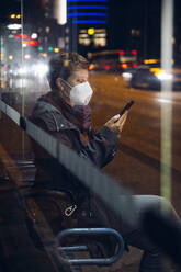 Mature woman wearing protective face mask using mobile phone while sitting at bus stop in city - MAMF01485