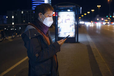 Frau mit Gesichtsmaske, die ein Mobiltelefon benutzt, während sie an einer Bushaltestelle in der Stadt steht - MAMF01486