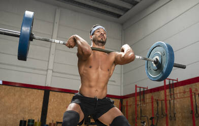 From below of concentrated male athlete lifting heavy barbell during weightlifting workout in gym while looking away - ADSF19594