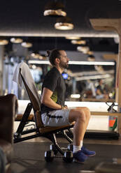 Seitenansicht eines erwachsenen Sportlers, der während einer Pause in einem modernen Fitnessstudio auf einer Bank sitzt - ADSF19601