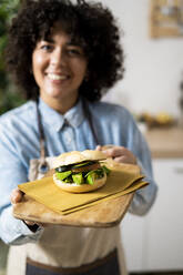 Junge Frau mit frisch zubereitetem veganem Sandwich mit Gurke, Avocado und Tomate - GIOF10363