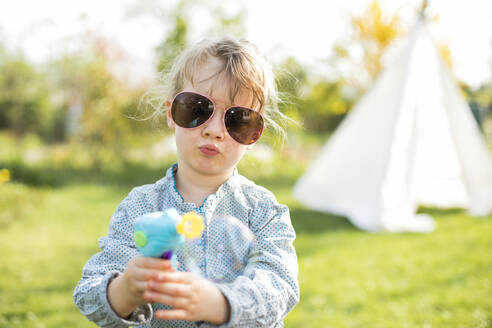 Mädchen mit Sonnenbrille, das mit einer Seifenblasenpistole spielt, während es im Garten steht - KVF00144