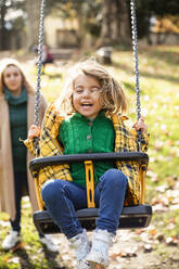 Lächelndes Mädchen, das auf einer Schaukel spielt, mit einer im Hintergrund stehenden Frau im Park - MCVF00680