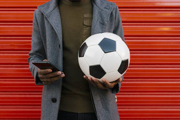 Man wearing jacket holding football while using mobile phone standing against red wall - EGAF01322