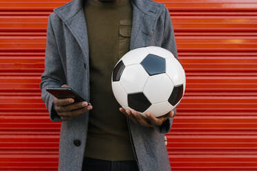 Mann mit Jacke, der einen Fußball hält, während er ein Mobiltelefon benutzt, steht vor einer roten Wand - EGAF01322