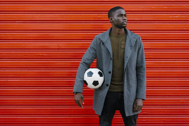 Young man with soccer ball looking away while standing against red wall - EGAF01329