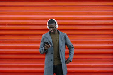 Lächelnder Mann mit Kopfhörern, der ein Mobiltelefon benutzt, während er an einer roten Wand steht - EGAF01334