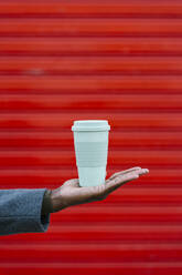 Man holding disposable cup in hand against red wall - EGAF01338