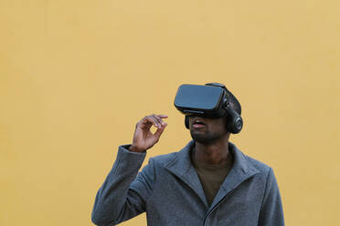 Young man using virtual reality headset while standing against yellow wall - EGAF01346