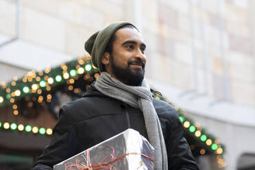 Lächelnder junger Mann mit Weihnachtsgeschenk in der Hand, während er an einem Gebäude in der Stadt steht - SGF02729