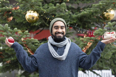 Lächelnder junger Mann, der ein Geschenk und ein Ornament vor einem Weihnachtsbaum hält - SGF02734