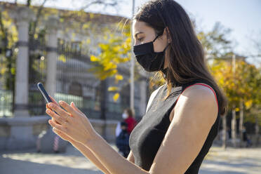 Junge Frau mit Gesichtsmaske, die ein Smartphone benutzt, während sie auf der Straße steht - IFRF00225