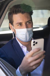 Close-up of businessman wearing mask using smart phone in taxi seen through window - IFRF00233