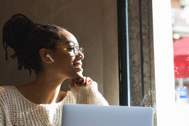 Lächelnde junge Frau, die durch das Fenster schaut, während sie mit ihrem Laptop in einem Café sitzt - PNAF00407
