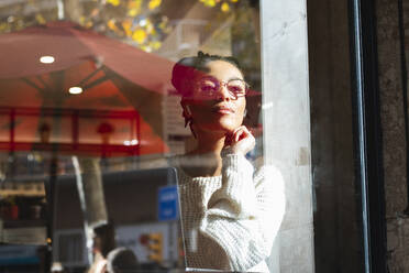 Selbstbewusste junge Frau mit Brille schaut durch das Fenster eines Cafés - PNAF00409