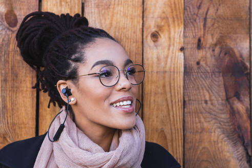 Lächelnde schöne junge Frau, die durch In-Ear-Kopfhörer gegen eine Holzwand spricht - PNAF00421