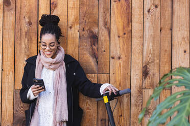 Schöne weibliche Millennial mit Smartphone durch elektrische Push-Roller gegen Holzwand - PNAF00422