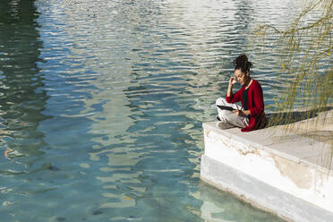 Junge Frau, die ein digitales Tablet benutzt, während sie im Schneidersitz am Rande der Promenade über dem Fluss an einem sonnigen Tag sitzt - PNAF00426