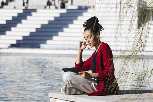 Junge Frau sitzt im Schneidersitz und benutzt ein digitales Tablet, während sie an einem sonnigen Tag am Rande einer Promenade über einem Fluss steht - PNAF00444