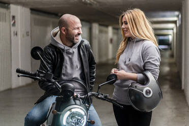 Lächelnde Frau im Gespräch mit ihrem Freund auf dem Motorrad sitzend auf einem Parkplatz - DLTSF01452