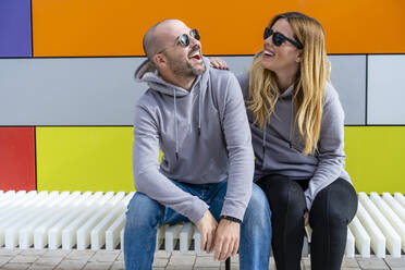 Fröhliches Paar mit Sonnenbrille sitzt auf einem Sitz vor einer bunten Wand - DLTSF01456