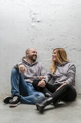 Glückliches Paar im Gespräch auf dem Fußweg vor einer weißen Wand sitzend - DLTSF01459