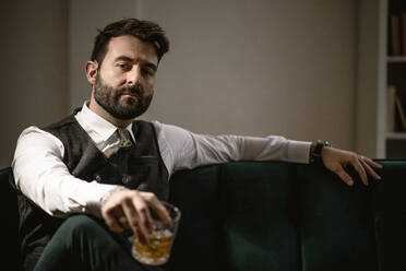 Porträt eines bärtigen Mannes auf dem Sofa mit einem Glas Whiskey in der Hand - GIOF10374