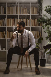 Portrait of bearded man sitting on stool and smoking pipe - GIOF10399