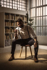 Portrait of bearded man sitting on stool and smoking cigar - GIOF10411