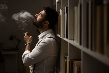 Porträt eines bärtigen Mannes, der vor einem Bücherregal Zigarre raucht - GIOF10417