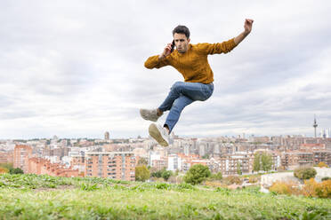 Young man jumping while on the phone over hill against cityscape - GGGF00577