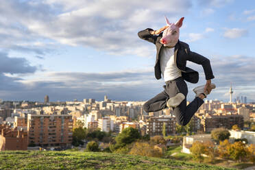 Geschäftsmann mit Schweinemaske springt auf Hügel in Stadt gegen Himmel - GGGF00585