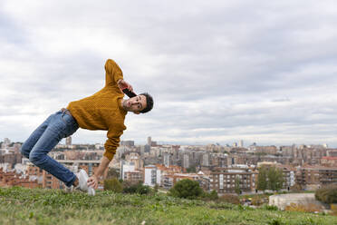 Junger Mann, der mit seinem Handy telefoniert, während er vor einer Stadtlandschaft akrobatische Übungen macht - GGGF00594
