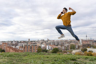 Excited young man talking on mobile phone while jumping over hill against cityscape - GGGF00600