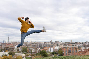 Excited man talking on mobile phone while jumping over hill against cityscape - GGGF00603