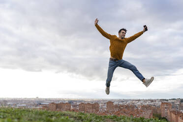 Excited young man holding mobile phone while jumping on hill against cityscape - GGGF00606