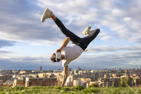 Flexible young man wearing virtual reality headset while doing handstand on hill in city against sky - GGGF00616