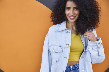 Smiling young woman with curly hair standing against heart shape on wall - SUF00646