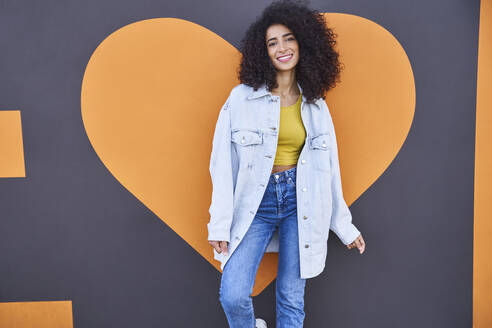 Happy young woman with curly hair standing against heart shape on wall - SUF00649